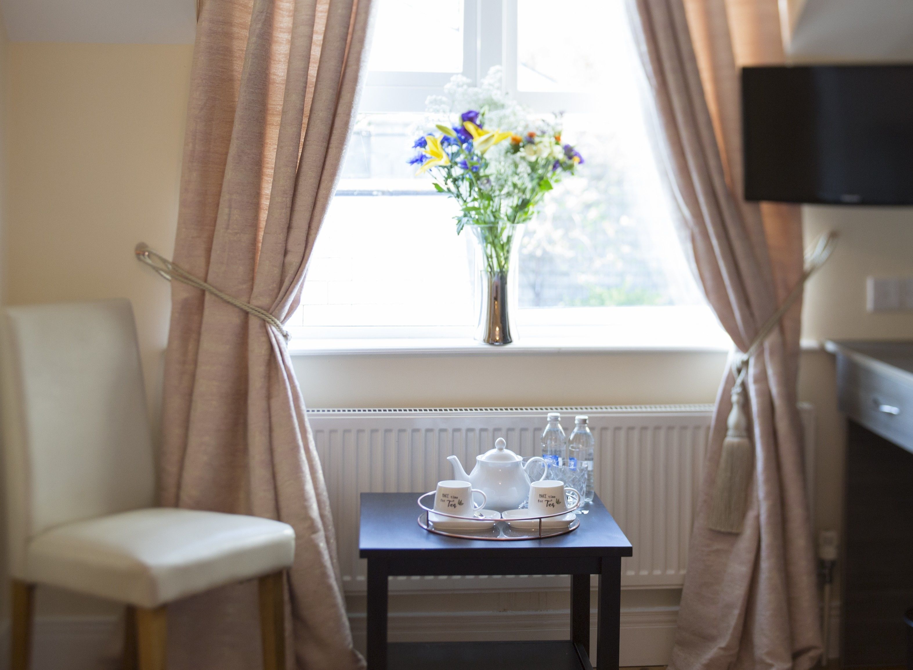 A room with a window, curtains, a chair, a table with a tea set, and a vase with flowers.