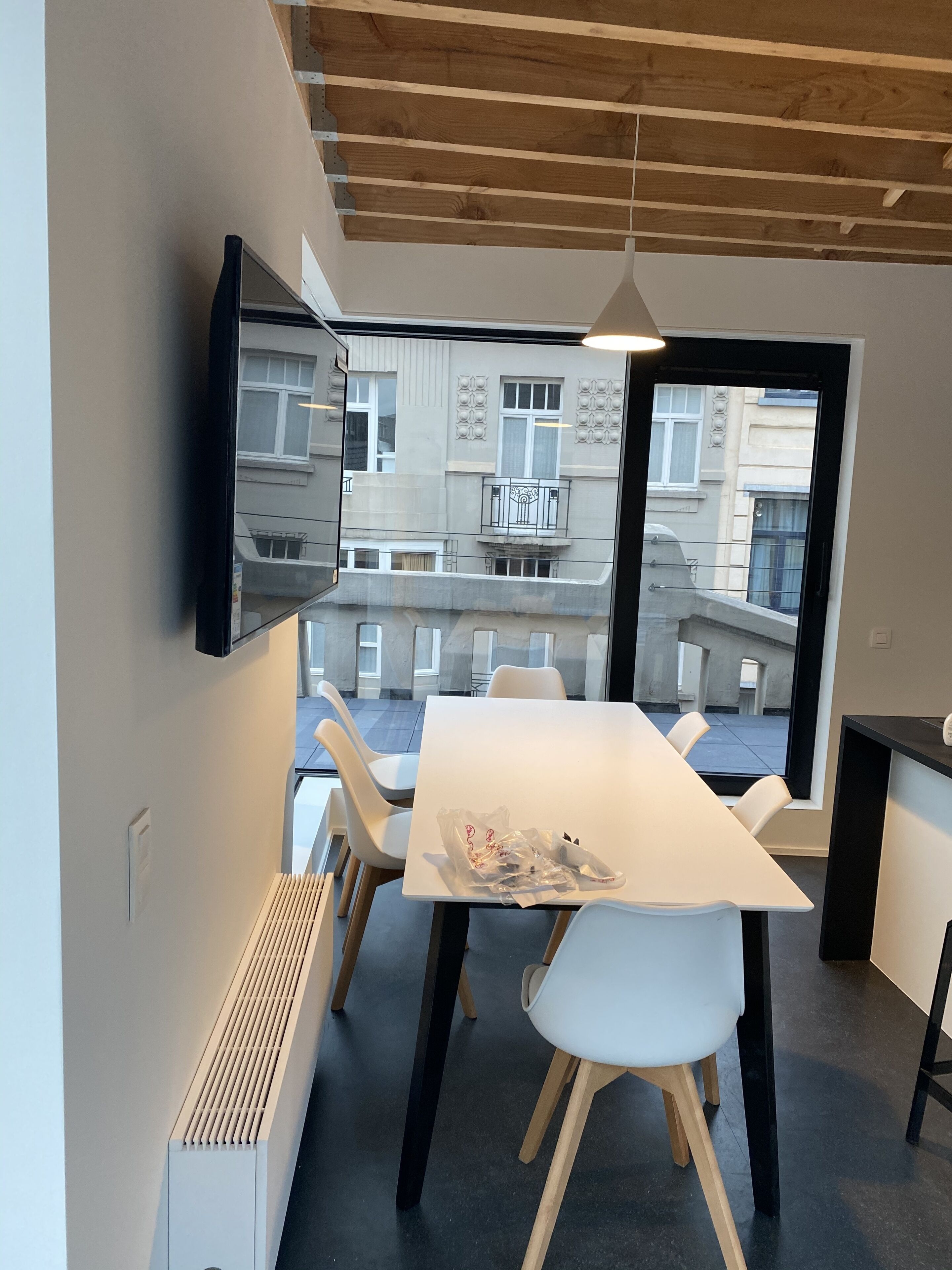 Un espace de repas moderne avec une longue table et des chaises blanches, un téléviseur fixé au mur et une vue sur l’extérieur du bâtiment à travers de grandes fenêtres.
