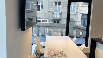 Un espace de repas moderne avec une longue table et des chaises blanches, un téléviseur fixé au mur et une vue sur l’extérieur du bâtiment à travers de grandes fenêtres.
