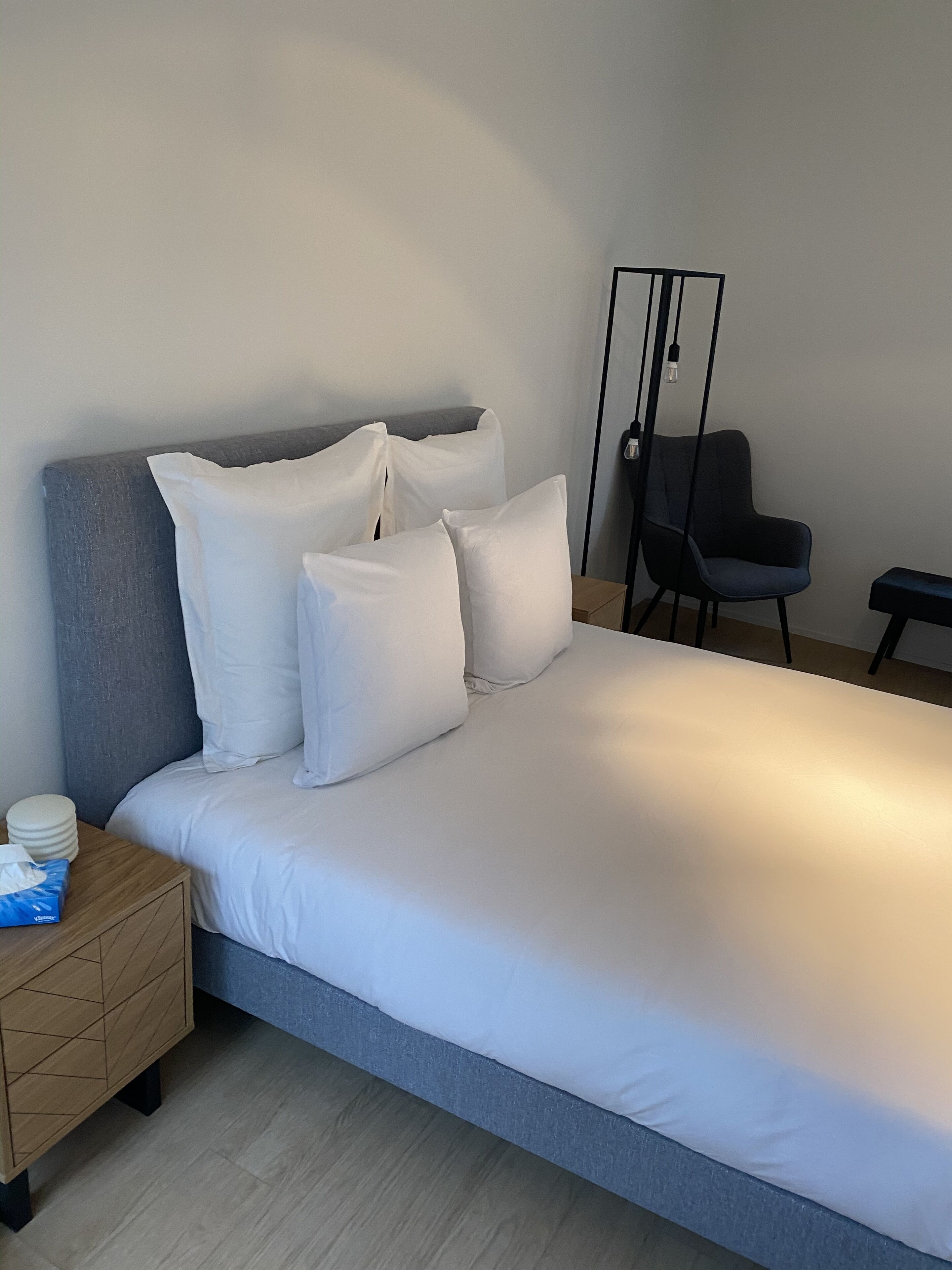 Un lit bien fait, avec du linge de lit blanc, une table de chevet en bois sur laquelle se trouve une boîte de mouchoirs, et une chaise moderne à la structure noire.