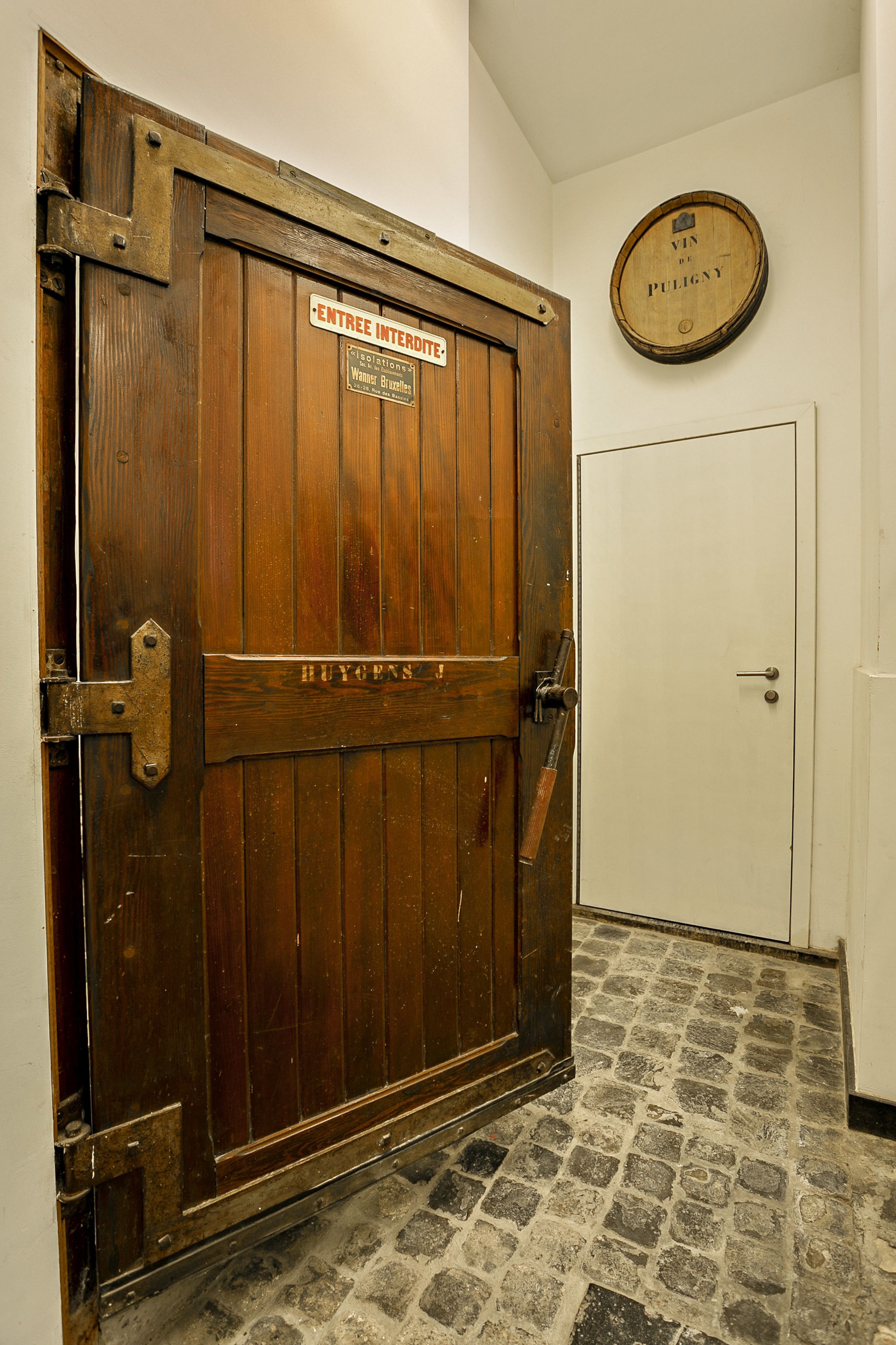 Una puerta de madera con un letrero que dice "ENTREE INTERDITE" y "HUGHES J." Una placa de madera redonda con "VIN DE PULIGNY" está montada en la pared sobre la puerta.