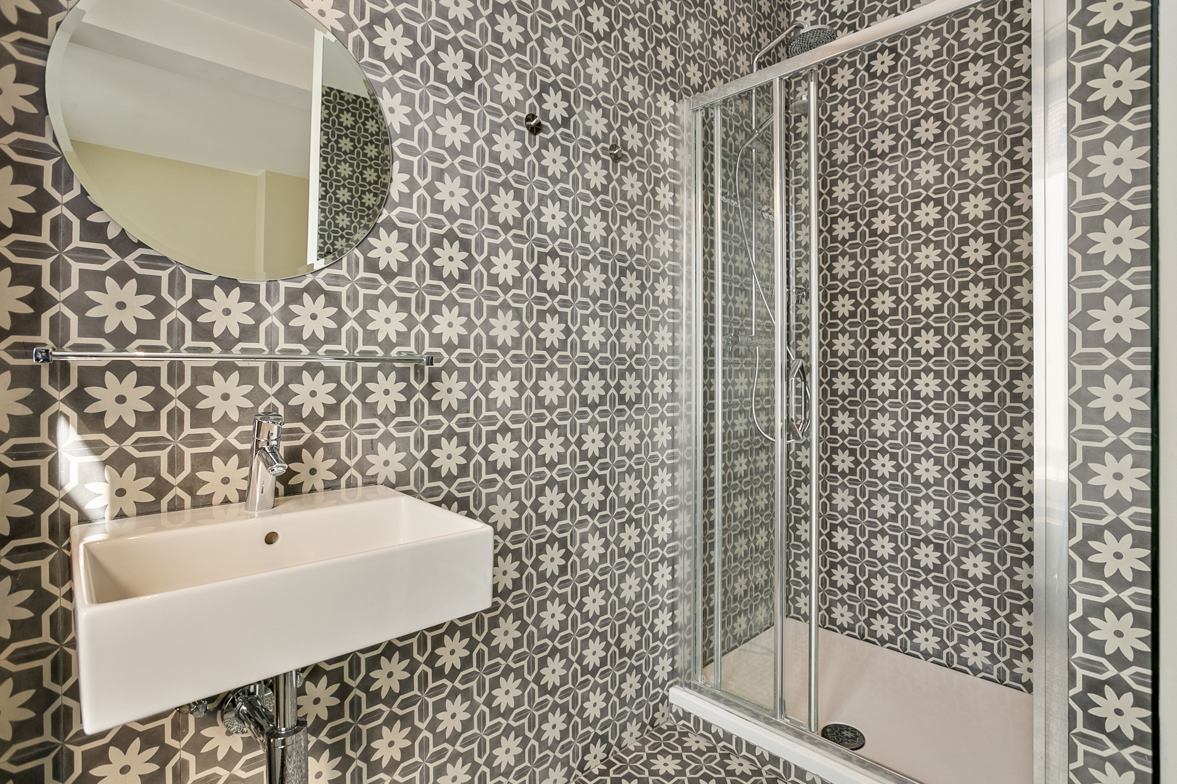 Un baño con lavamanos, espejo y ducha con azulejos estampados.
