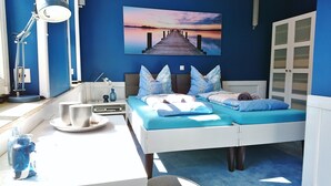 A bedroom with a bed, pillows, a bedside table, a lamp, a painting, and a view of the sea.