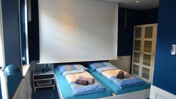 Una habitación con dos camas, un televisor blanco montado en el techo, una mesita con lámpara y un mueble con puertas de vidrio satinado.