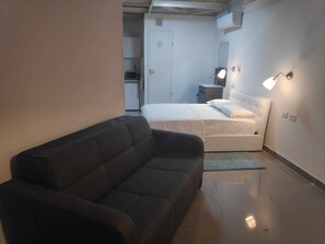 A compact hotel room with a dark-colored sofa, a bed with white bedding, a small bedside table, and a wall-mounted lamp.