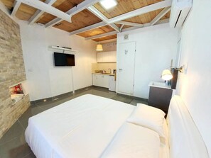 A bedroom with a large bed, a TV mounted on the wall, a kitchen area, and a wooden ceiling.