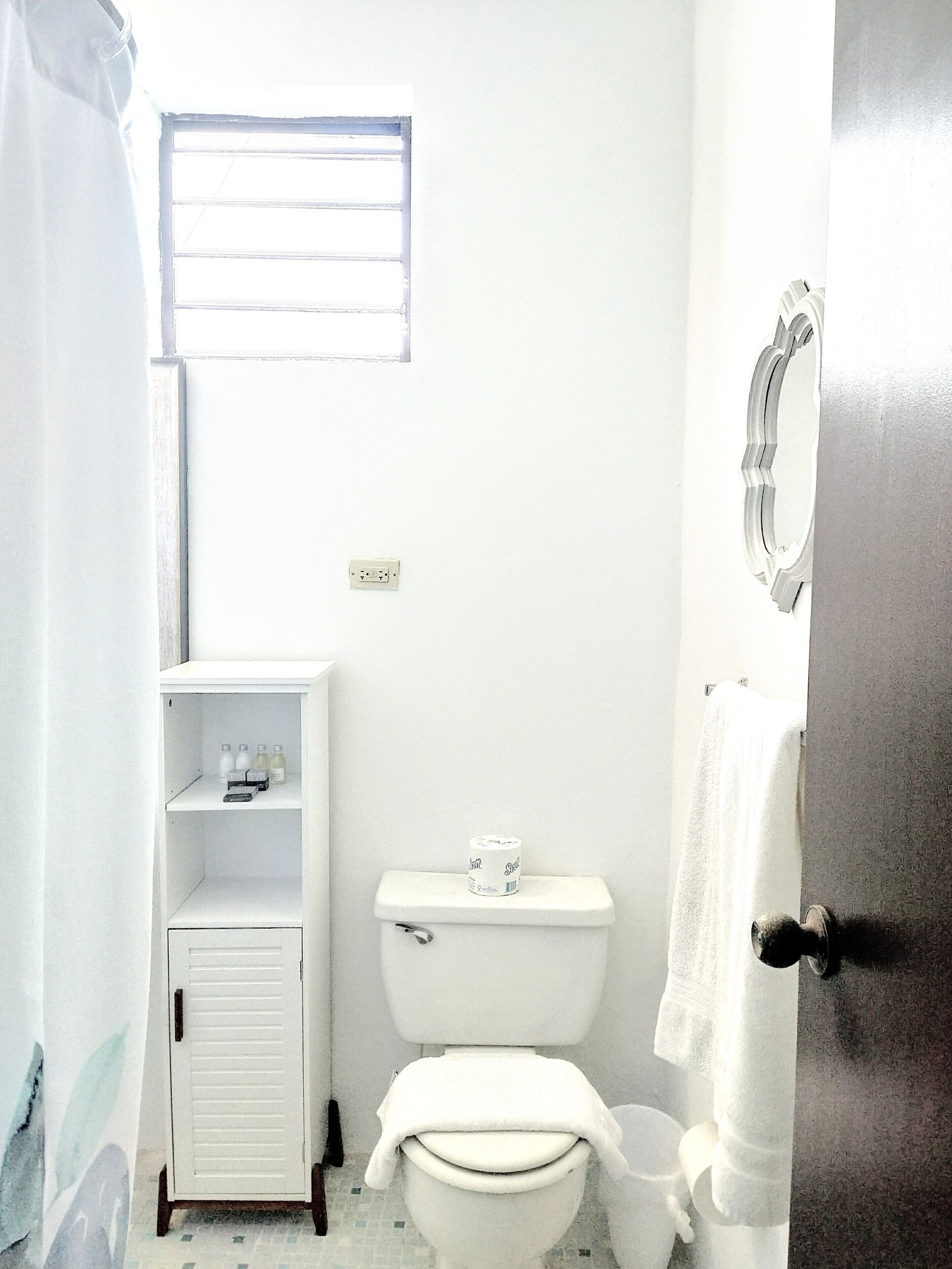 Et lille, hvidt badeværelse med toilet, et hyldesæt med toiletartikler og et vindue over toilettet.
