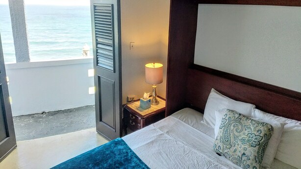 Ein Hotelzimmer mit einem Bett, einem Nachttisch, einer Lampe und Blick auf das Meer durch eine offene Tür.
