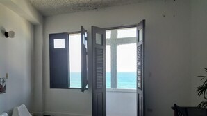 Ein Zimmer mit Schachbrettmuster-Boden, zwei weiße Plastikstühle und einem Holztisch, mit Blick auf das Meer durch eine geöffnete Tür.
