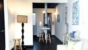 Ein modernes Wohnzimmer mit einem weißen Sofa, einem Couchtisch und zwei Holzschemeln. Im Hintergrund befindet sich ein Essbereich mit Tisch und Stühlen.