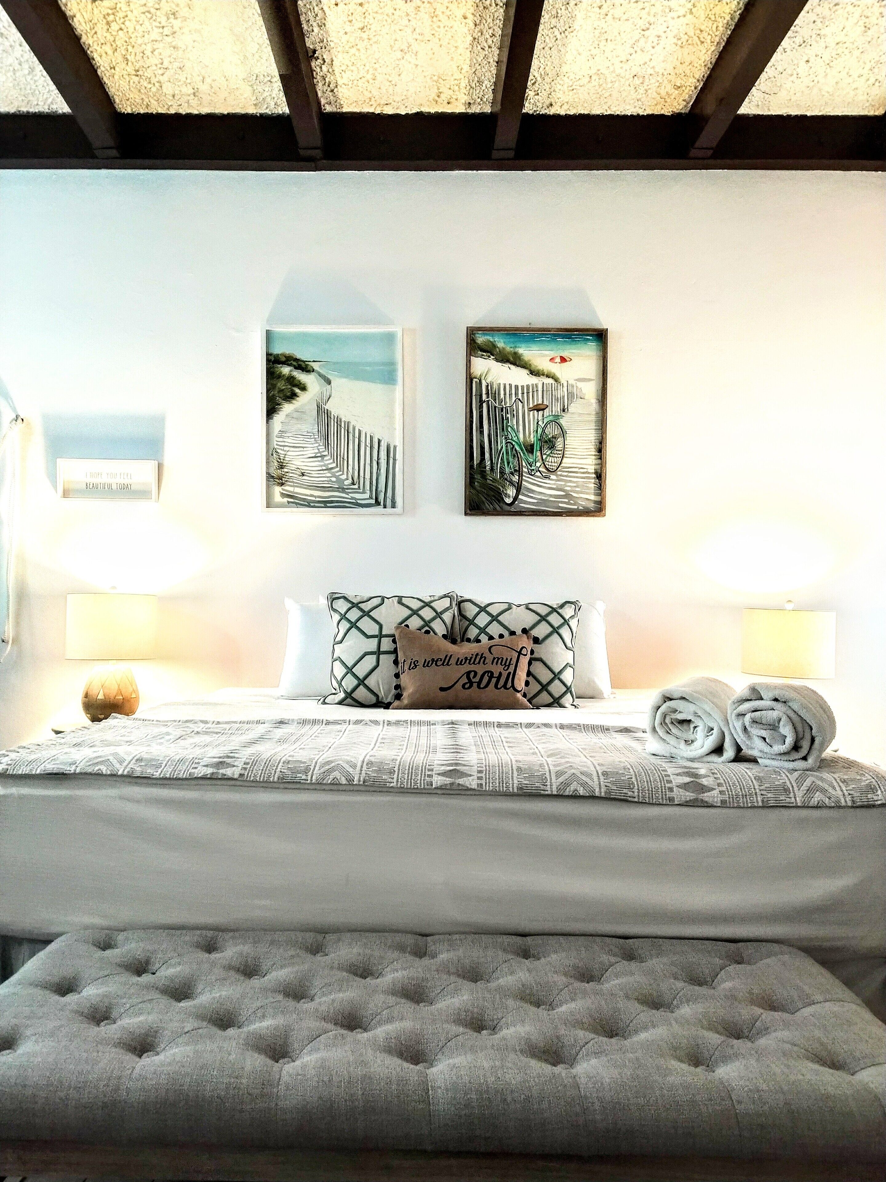 Un dormitorio con una cama, dos obras de arte colgadas en la pared que representan una escena de playa, un cojín con una cita y una lámpara de noche.
