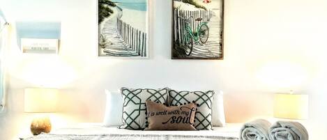 Un dormitorio con una cama, dos obras de arte colgadas en la pared que representan una escena de playa, un cojín con una cita y una lámpara de noche.