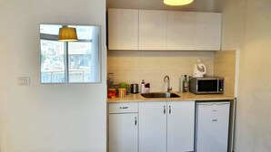 A compact kitchen with white cabinets, a microwave, a sink, and a dishwashing unit. There is a window with a view of buildings outside. - Agas Holiday Apartments Gefen Boutique (Tiberias)