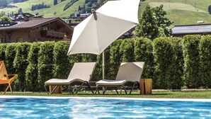 A pool area with two lounge chairs and a large umbrella, surrounded by neatly trimmed hedges and a view of a mountainous landscape. - Rosentalerhof Hotel & Apartments (Saalbach-Hinterglemm)