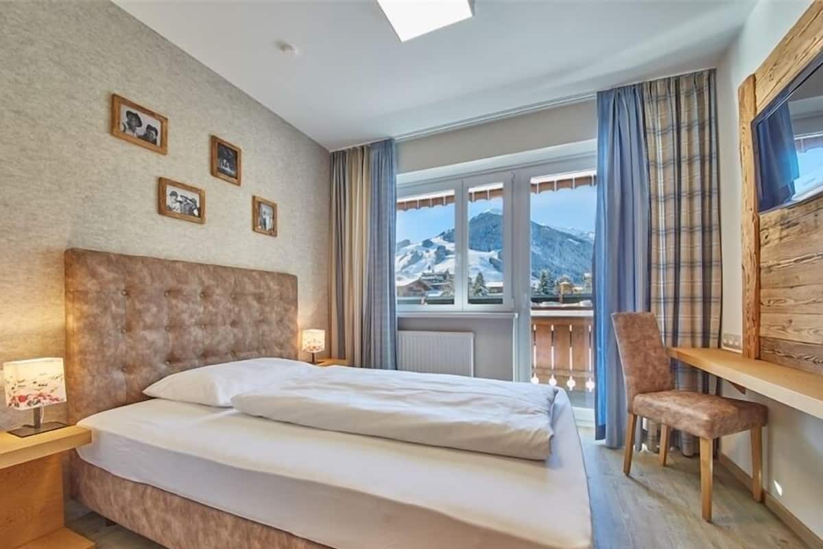 A hotel room with a large bed, a wooden desk, a chair, and a view of mountains.