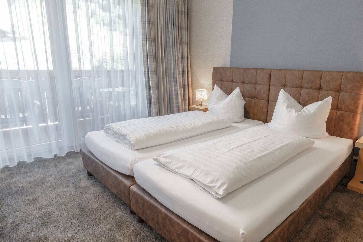 A double bed with a wooden headboard, two bedside tables with lamps, sheer curtains, and a carpeted floor.