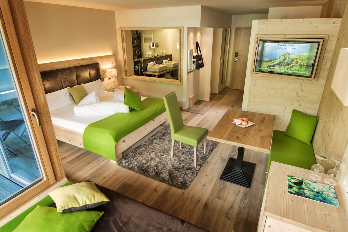 Une chambre d’hôtel moderne comprenant un lit, des sièges recouverts d’un tissu vert, un coin repas, un téléviseur et une vue sur la pièce.
