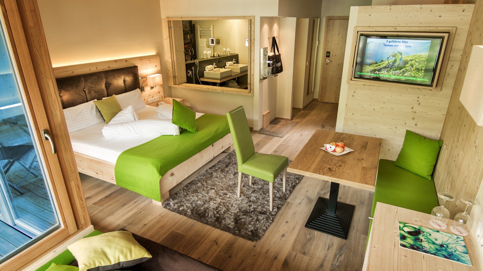 Habitación de hotel moderna con cama, escritorio, silla, televisor y baño visible a través de un espejo.