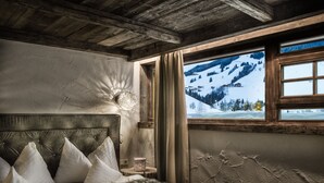 Una habitación con techo de madera, cama con almohadas blancas, manta de piel y vista a un paisaje montañoso cubierto de nieve a través de una ventana.