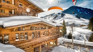 A wooden chalet with a ski slope in the background. - Wellnesshotel Alpin Juwel (Saalbach-Hinterglemm)
