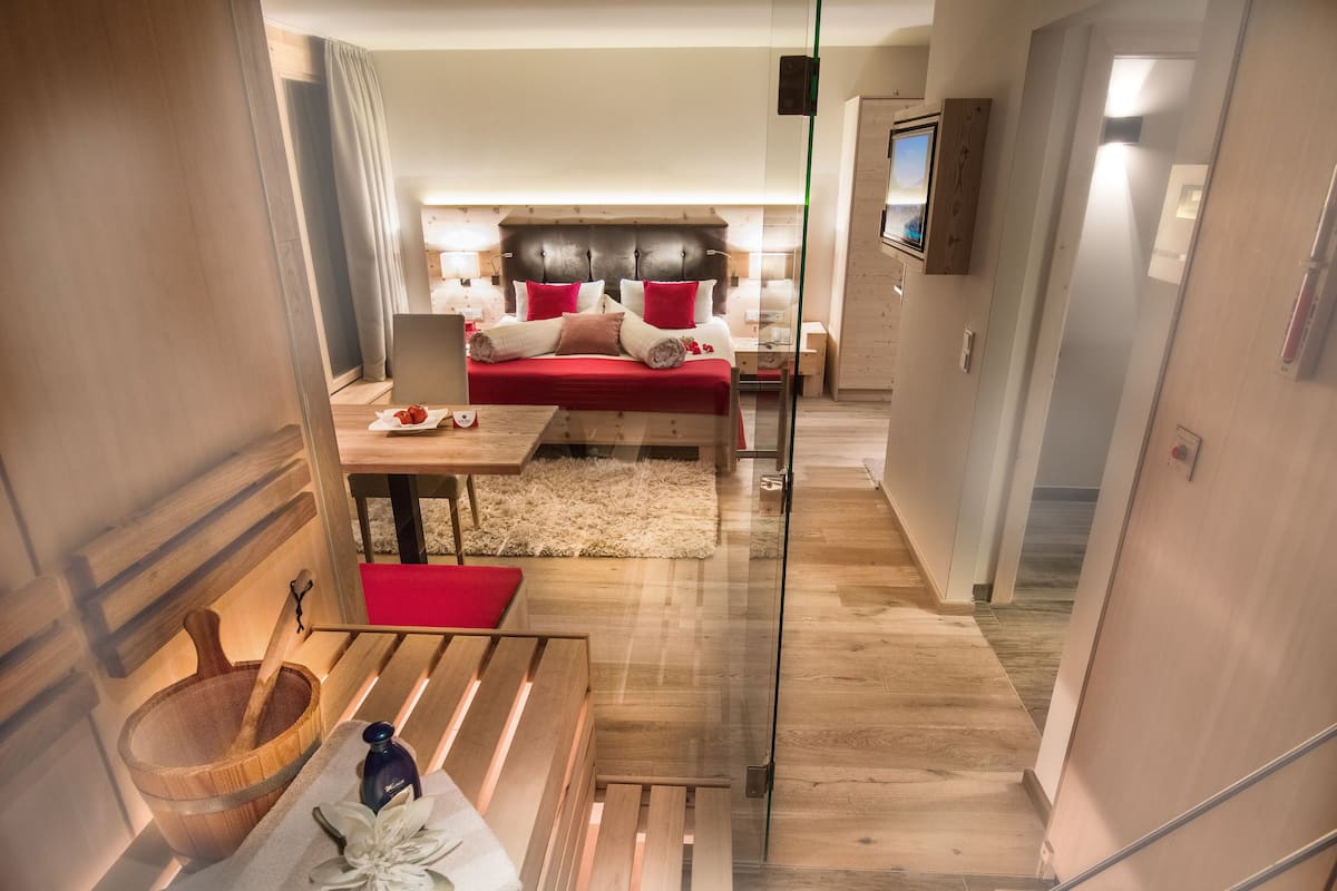 Une chambre d’hôtel moderne avec un plancher en bois, un lit avec une literie rouge et blanche, une petite table avec une bouteille et des fleurs, et un téléviseur fixé au mur.