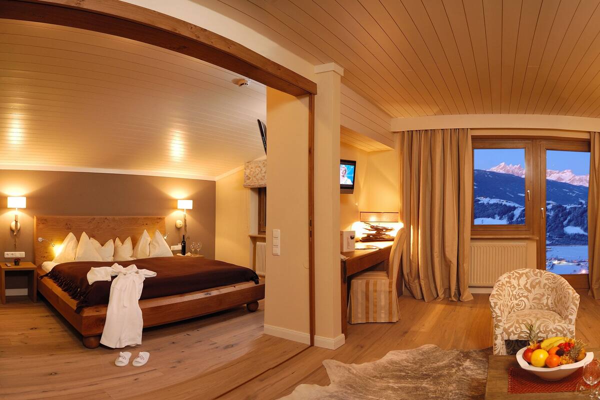Une chambre d’hôtel avec un lit, un bureau, une chaise, un canapé, un panier de fruits et une vue sur les montagnes par la fenêtre.