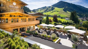 A hotel with a wooden facade, outdoor seating with umbrellas, and a view of green hills and mountains. - Wellnesshotel Alpin Juwel (Saalbach-Hinterglemm)