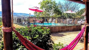 A hammock tied between wooden posts, a pool area with lounge chairs and umbrellas, and a view of hills in the background. - La Loma Hotel Barichara (Barichara)