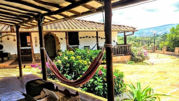 A covered outdoor area with a hammock, a table with a basket, and a view of a building with a tiled roof and a garden with various plants. - La Loma Hotel Barichara (Barichara)