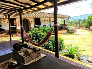 Un espacio al aire libre techado con una hamaca, una mesa con una cesta, y vista a un edificio con techo de tejas y un jardín con diversas plantas.
