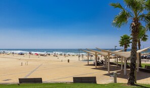A beach with a covered seating area, palm trees, and a clear blue sky. - Lagoon Amazing water front 3 (Herzliya)