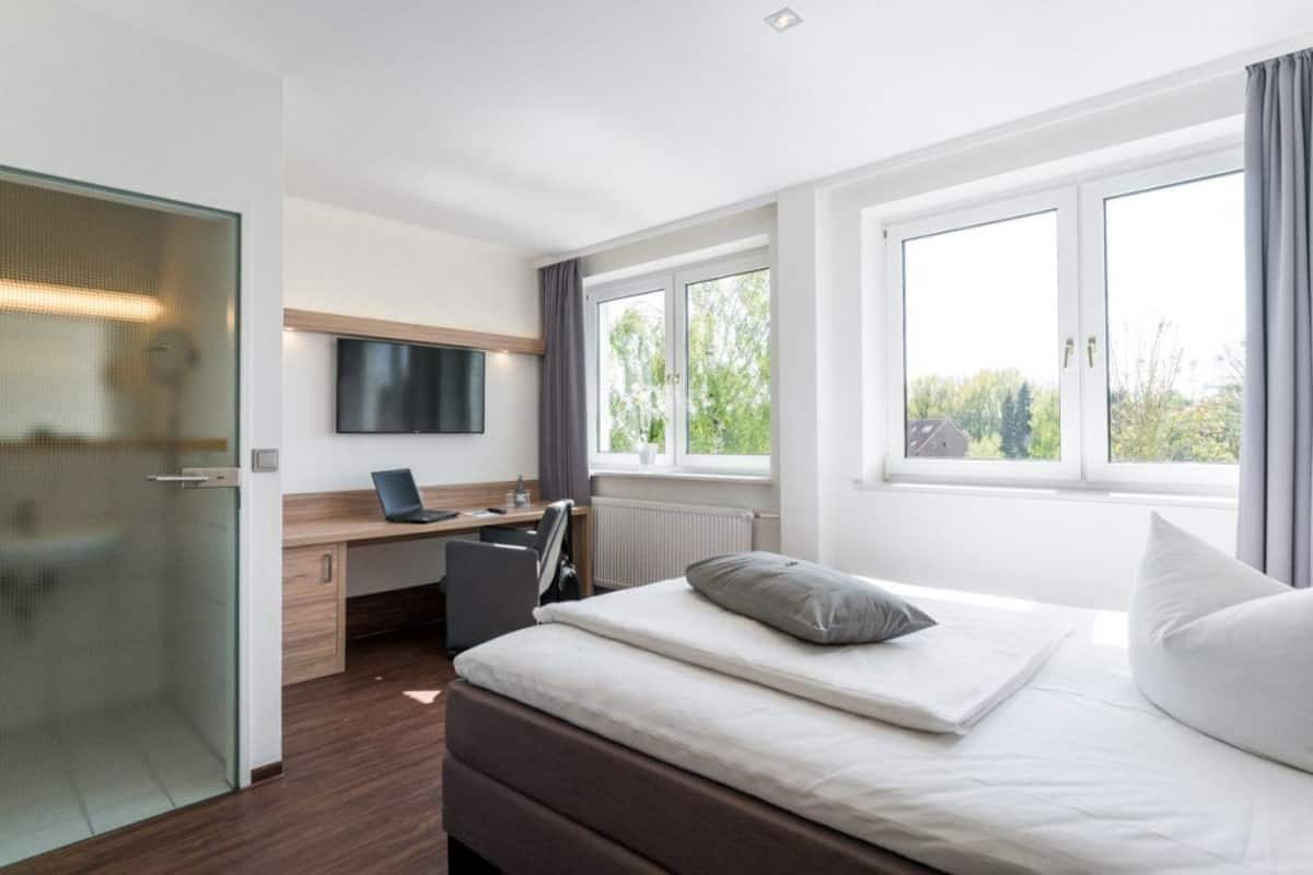A hotel room with a bed, a desk with a laptop, a TV, and a window with a view of greenery.