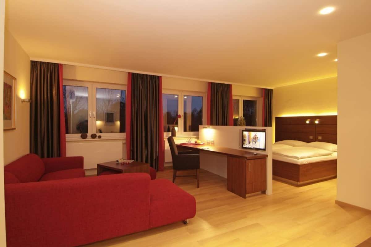 A hotel room with a red sofa, a wooden desk, a bed, and large windows with curtains.