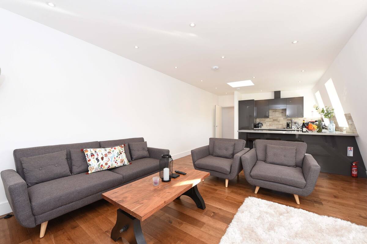 Ein modernes Wohnzimmer mit einem grauen Sofa, zwei Sesseln, einem Holztisch und einer Küche im Hintergrund.