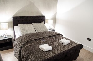 A neatly made bed with a dark headboard, two pillows, and two rolled towels on the bed. There are two bedside tables with lamps and a small decorative item on each. The room has a clean, modern look with light-colored walls and wooden flooring.