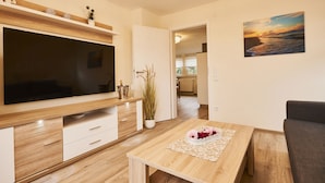 A modern living room with a flat-screen TV, wooden furniture, a dining table with a placemat, and a painting of a sunset on the wall.