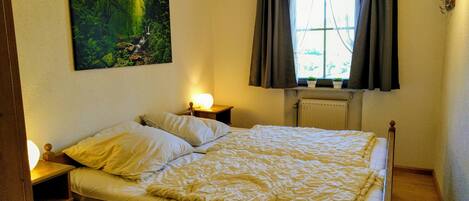 A bedroom with a bed, bedside lamps, a radiator, and a painting of a forest scene.
