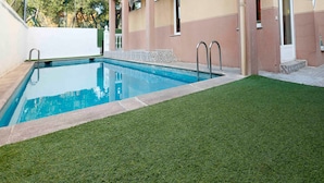 Una piscina con un pequeño área verde frente a ella, rodeada por un edificio de fachada beige y rosa.
