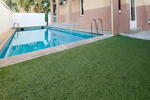Una piscina con un pequeño área verde frente a ella, rodeada por un edificio de fachada beige y rosa.