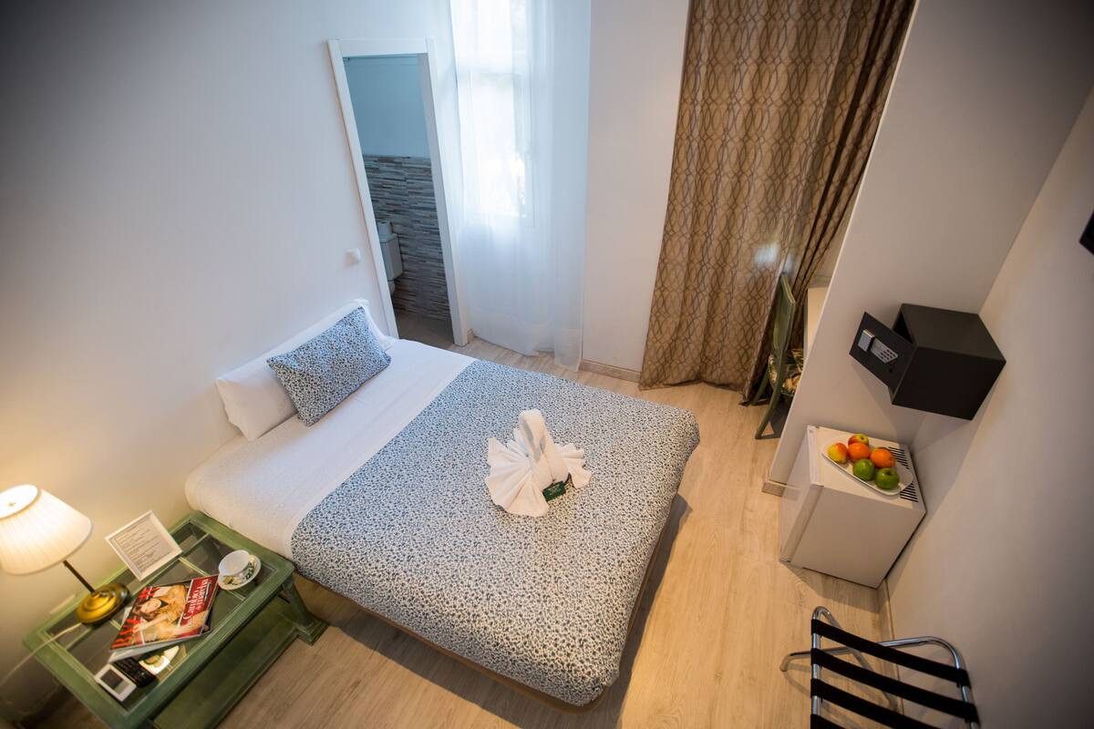 A hotel room with a bed, a bedside table with a lamp, a small table with fruits, and a chair.