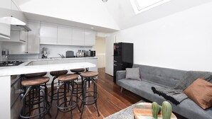 Eine moderne Küche mit weißen Schränken, einer zentralen Kochinsel mit Hockern und einem grauen Sofa mit Kissen.