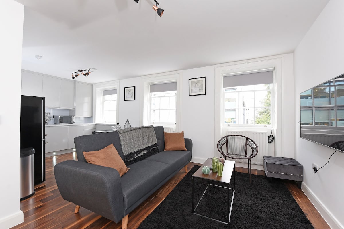 Una sala de estar moderna con un sofá gris, un tapete negro y una mesa de centro. Se ve una cocina al fondo con armarios blancos y un refrigerador negro.