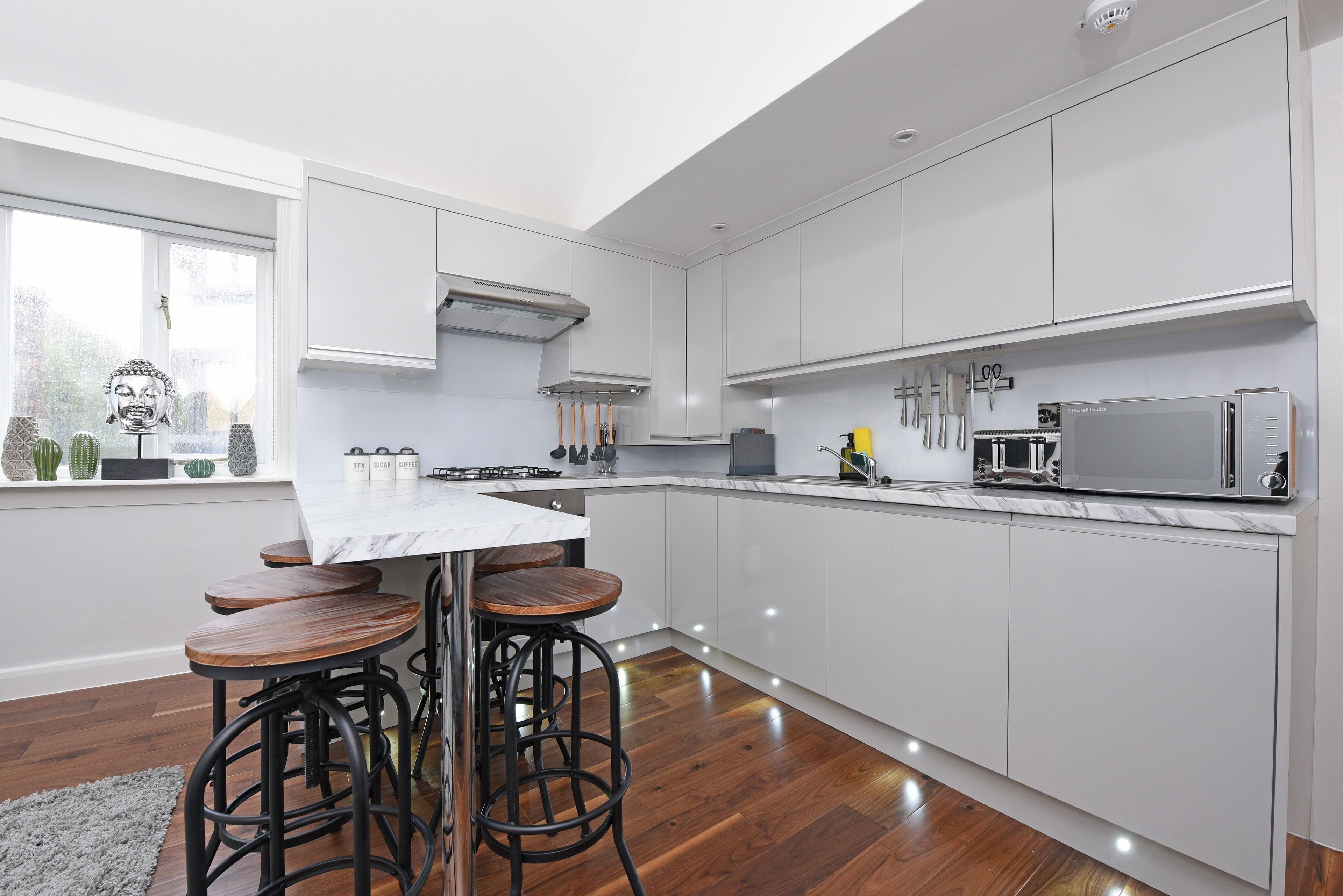 A modern kitchen with white cabinets, a marble countertop, and a built-in oven. There are three wooden stools and a window with a view of the outside.