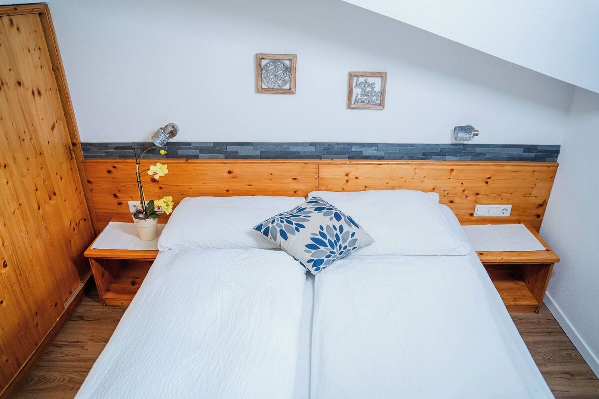 A bedroom with a wooden headboard, a bed with white bedding, two bedside tables, a nightstand, a potted plant, and two wall decorations.