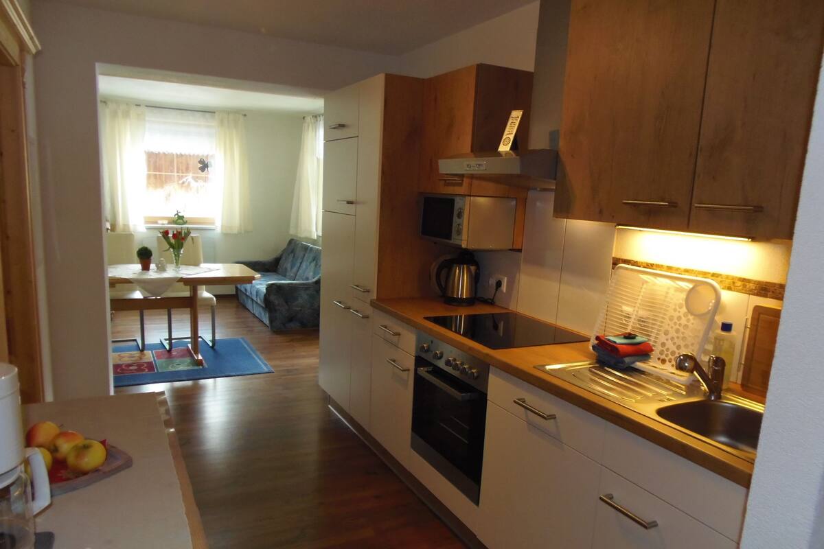 A compact kitchen with wooden cabinets, a microwave, an oven, and a sink. There is a dining area with a table and chairs visible in the background.