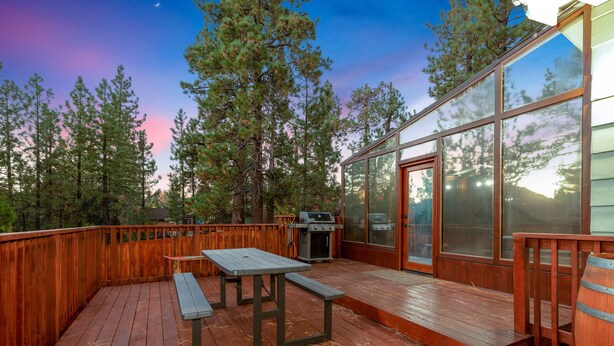 A wooden deck with a grill and a table, surrounded by tall trees and a glass door leading to a building.