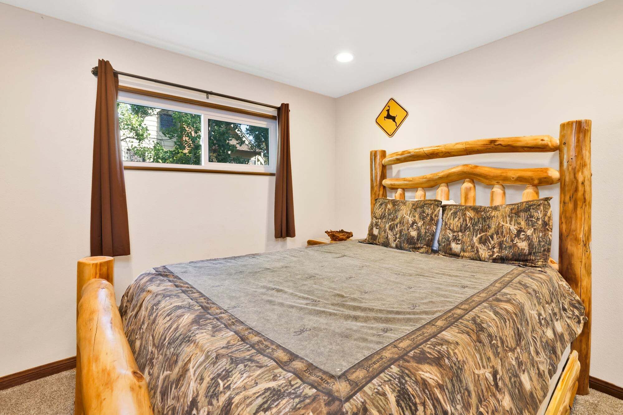 Een slaapkamer met een houten bed, een raam met gordijnen, een waarschuwingsbord met een hert en een tafeltje met een kom.