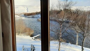 A view through a window of a river with snow-covered banks and trees, a bicycle leaning against the window frame, and a clear sky. - Guesthouse Hagi 1 (Husavik)