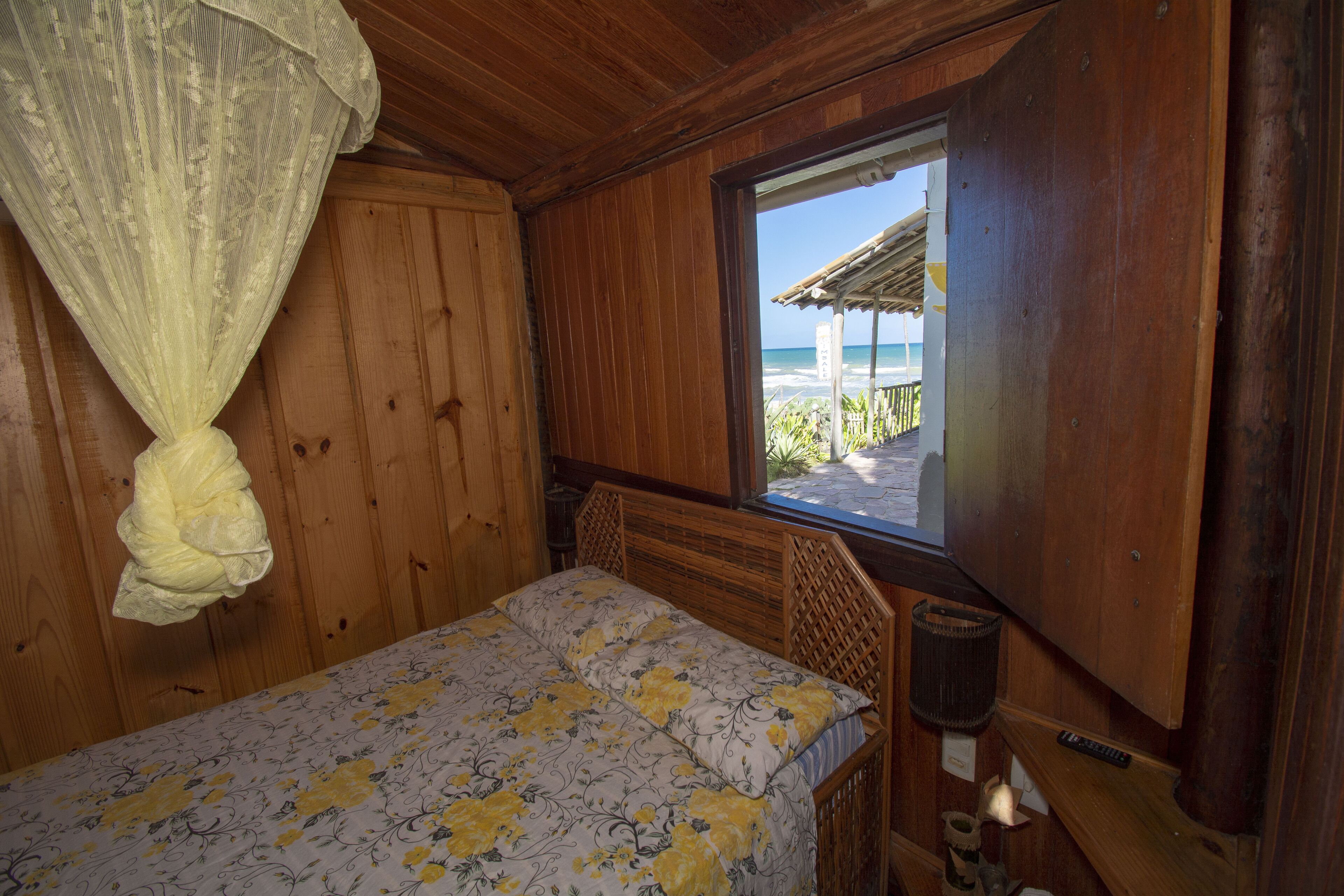 Una habitación de madera con una cama, un ventanal con vistas a la playa y una mosquitera.
