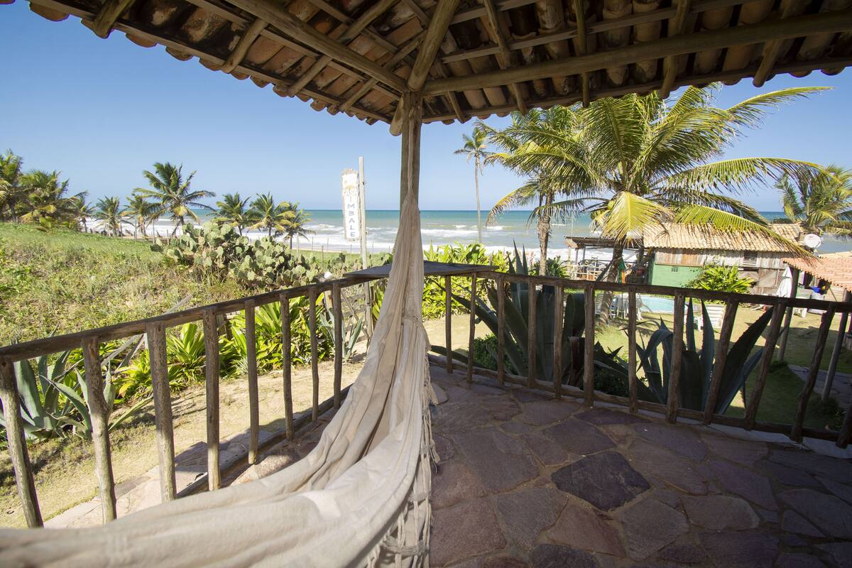 A covered outdoor area with a hammock, palm trees, and a view of the beach and ocean.